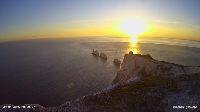 A view of white chalk cliffs extending into the ocean at sunset, with a few prominent rocks in the water near the cliffs. The sky is clear, and the sun is low on the horizon, casting a golden reflection on the sea. The timestamp in the bottom left corner reads "29/04/2025 20:00:07," and the website "isleofwight.com" is displayed in the bottom right corner.