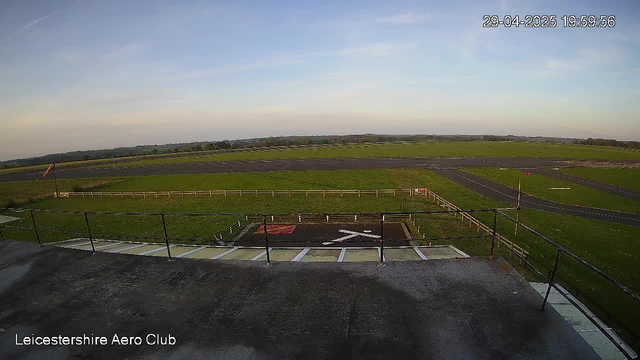 An empty aircraft tarmac area with a small, closed helipad marked with an "X" in red paint in the foreground. Surrounding the tarmac is a low fence, and beyond that, a vast grassy field stretches into the distance. There are some small roadways and pathways visible, along with a few signs and posts. The sky is mostly clear with some light clouds, and the time stamp indicates late evening. The bottom left corner has the text "Leicestershire Aero Club."