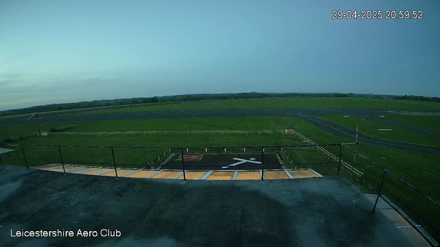 An empty helicopter landing pad with a white helicopter symbol in the center, surrounded by green grass fields extending to the horizon. The sky is partly cloudy with a transition from light to darker blue, indicating late evening. The image has a timestamp in the top right corner and the words "Leicestershire Aero Club" in the bottom left corner.