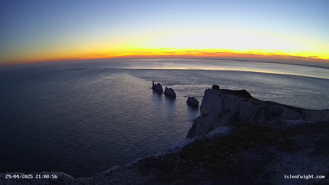 A view of a coastal scene at sunset or sunrise, with the sky displaying a gradient of colors from deep blue to warm orange and yellow. The ocean stretches out to the horizon with gentle waves, and there are several large rock formations rising from the water near a white chalk cliff. A small lighthouse or navigation tower is visible on one of the rocks, emitting a faint light. The foreground shows the edge of the chalk cliff with patches of grass and dark soil. The timestamp in the bottom left corner indicates the date and time as April 29, 2025, at 21:00:56, and the bottom right corner displays the website "isleofwight.com".