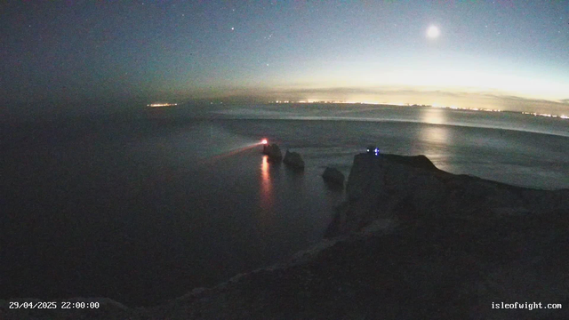 A nighttime scene of a calm ocean with a rocky shoreline in the foreground. The sky is dark with a few visible stars and a bright, nearly full moon casting reflections on the water. Distant city lights are faintly visible on the horizon. A faint light source is seen among the rocks near the water's edge, and the scene appears tranquil and serene.