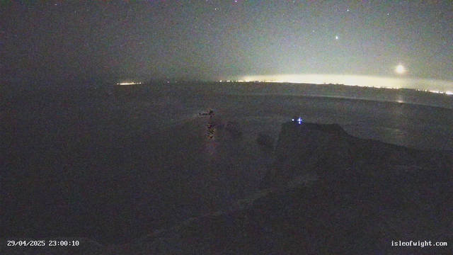 A night-time scene of a beach with dark sand and water, illuminated faintly by the moon and distant city lights on the horizon. The sky is clear, showing stars, and there are no visible people or animals. The timestamp indicates it is late at night on April 29, 2025.