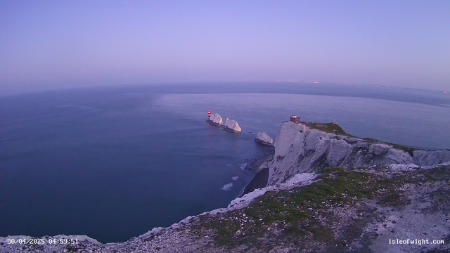 A view of white chalk cliffs over the ocean at dusk or dawn, with three large rock stacks near the shore. A lighthouse with a red and white tower is visible on one of the rocks. The sky has a gradient of soft pastel colors, transitioning from purple to pink. The scene is calm and serene, with gentle waves lapping against the rocks.