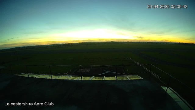 A sunrise over an open grassy airfield with a clear sky, showing the distant horizon with a few clouds and a faint orange glow from the rising sun. The image is taken from a webcam at Leicesterhire Aero Club, dated April 30, 2025, at 5:00 AM. The foreground features a helipad with a helicopter landing mark, surrounded by a railing.
