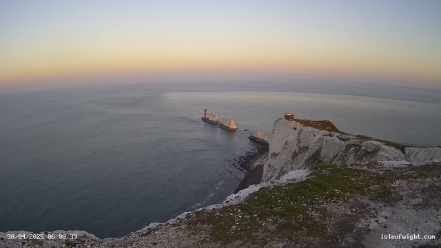 A view of white chalk cliffs and rock formations along a coastline at sunrise, with the calm sea stretching to the horizon. There is a lighthouse mounted on one of the rocks and a small building or structure on the top of the highest part of the cliff. The sky displays a gradient of soft pastel colors, transitioning from pale yellow near the horizon to light blue higher up.