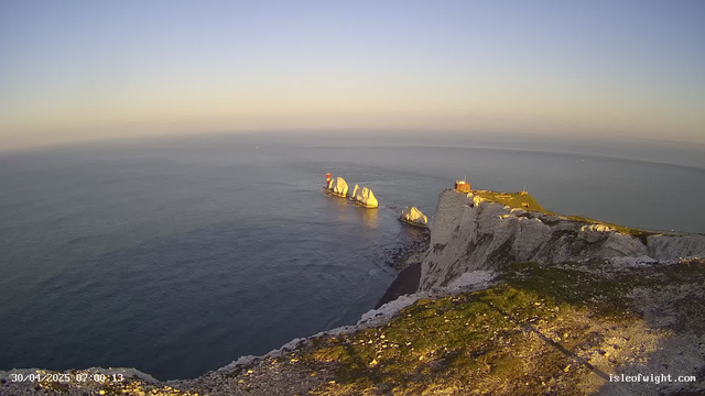 A panoramic view of high white chalk cliffs overlooking the ocean during sunrise or sunset. A lighthouse and some buildings are situated on the edge of the cliffs, with a grassy area surrounding them. Several large rocks are visible in the water just offshore. The sky is clear with a gentle gradient from light to darker blue, and the sea appears calm with subtle waves. The timestamp in the lower left corner indicates the photo was taken on April 30, 2025, at 07:00:13.
