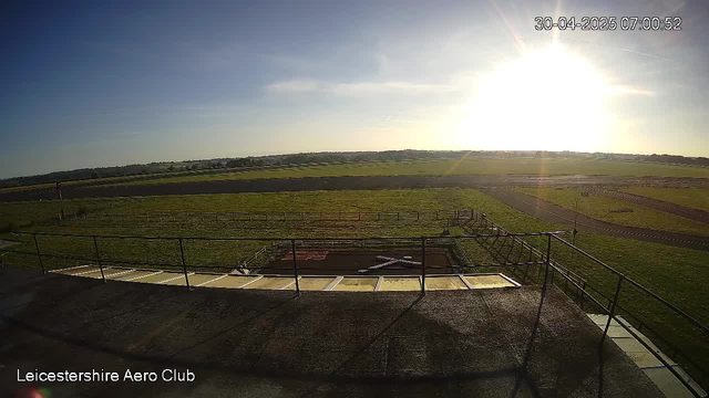An outdoor scene with a clear sky and bright sunlight, likely in the early morning. The image shows a grassy field with fences and paved pathways stretching into the distance. The view is from a high vantage point, possibly from a control tower or similar structure. The timestamp reads "30-04-2025 07:00:52," and the bottom left corner has text that says "Leicestershire Aero Club."
