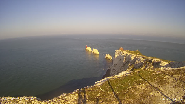 A panoramic view of white chalk cliffs overlooking the ocean with a lighthouse and a small building on top. Several large white rocks are visible in the water near the cliffs, and the sky is clear with a soft gradient from blue to light near the horizon. The time and date are displayed in the bottom left corner.