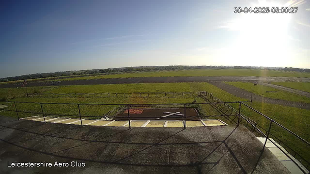 An outdoor view from a webcam showing a bright, sunny sky with some light clouds. The landscape features open grassy fields, a paved runway, and a fenced area in the foreground. The timestamp indicates the date as April 30, 2025, and the time as 08:00:27. The text "Leicestershire Aero Club" is visible in the lower left corner.