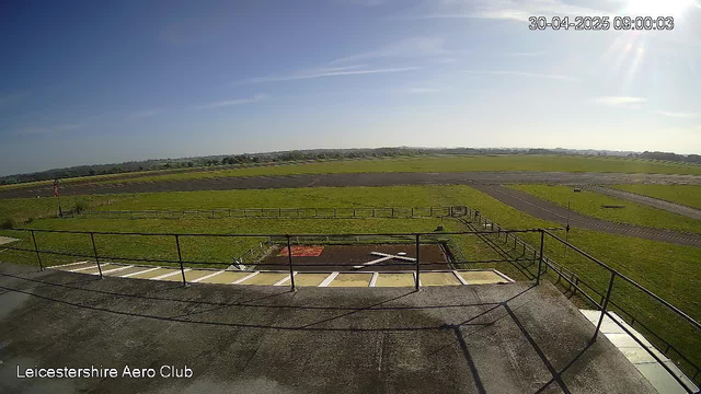 A wide outdoor view from a webcam at Leicestershire Aero Club, showing a sunny day with a clear blue sky, a grassy field with some paved pathways, and a distant horizon with low-lying hills. The foreground includes a concrete platform with metal railings, and there is a small red and black area on the ground directly below the camera. The timestamp on the image is April 30, 2025, at 9:00 AM.