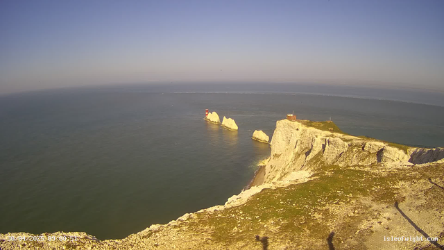 A scenic view of white chalk cliffs overlooking the sea, with a small lighthouse and a building on top of the cliffs. Large rock formations are visible in the water near the shore, and the sky is clear with soft lighting suggesting late afternoon or early evening. The horizon extends far into the distance, blending with the calm ocean.