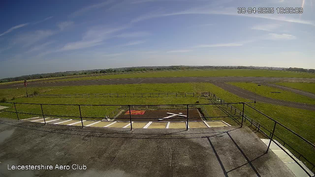 An outdoor scene showing a view from a webcam at LeicesterShire Aero Club. The image captures a clear sky with some wispy clouds, a grassy field, and paved pathways. The camera is positioned above a small parking area with numbered parking spots, railings, and a tarmac area. The timestamp in the upper right corner indicates the date as April 30, 2025, and the time as 10:00:09.