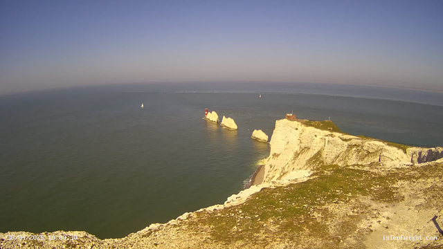 A panoramic view of white chalk cliffs along a coastline with a lighthouse and a small building on top, overlooking the ocean with sailboats in the distance on a clear day with blue skies.