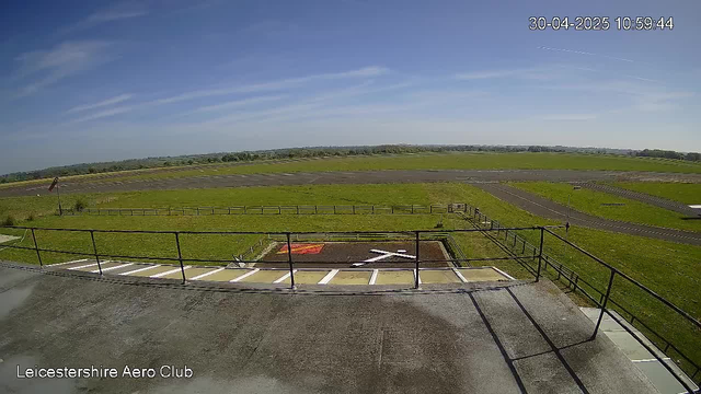 A wide view of an outdoor airfield at Leicestershire Aero Club under a clear blue sky with a few wispy clouds. The foreground shows a small parking area with marked parking spaces and a balcony with a black railing. There are grassy fields stretching into the distance, bordered by roads and pathways, with no aircraft visible. The timestamp in the top right corner reads "30-04-2025 10:59:44."