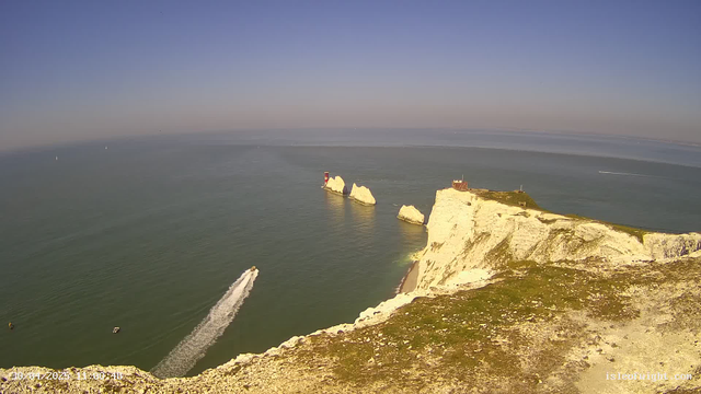 An aerial view of white chalk cliffs overlooking the sea, with a lighthouse and a small building on top of the cliffs. A boat is leaving a trail in the water below, and there are several sailboats farther out on the horizon. The sky is clear and blue.
