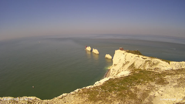 A view from a webcam showing white chalk cliffs extending into the ocean under a clear blue sky. There are several rock stacks near the base of the cliffs, with a lighthouse on one of the larger stacks. The water is calm, and a few boats are visible in the distance.