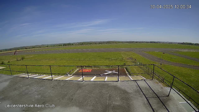 A view of an airport runway and grassy field taken from a webcam at Leicestershire Aero Club. The sky is clear with a few wispy clouds. There's a small helipad with a red and white marking visible in the foreground, and a fence surrounds the area. The date and time displayed in the upper right corner are 30-04-2025, 12:00:34.