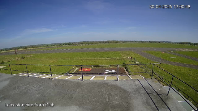 An outdoor view of a small airfield with a parking area and a helipad marked with a white "H". The sky is clear and blue with minimal clouds. In the distance, there are grassy open fields and a runway. The photo is timestamped "30-04-2025 13:00:48" and has the label "Leicestershire Aero Club" in the bottom left corner.