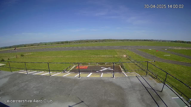 An outdoor view of a flight line at Leicestershire Aero Club on a clear day with blue skies. In the foreground, there is a concrete platform with a small helipad marked by a white 'H' and a red square. The platform is surrounded by black railings. Beyond the platform, there is a grassy field extending to the horizon, with a paved runway or taxiway running horizontally across the scene. The background consists of open green fields and distant trees, with a few small structures or objects barely visible in the distance. The date and time are displayed in the top right corner as 30-04-2025 14:00:02.