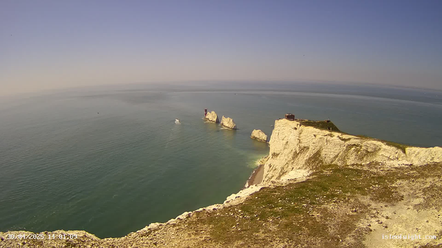 A panoramic view of white chalk cliffs overlooking the ocean, with several large rock formations protruding from the water near the shoreline. The sky is clear with a subtle gradient from light to deeper blue, indicating a calm and sunny day. A small building or structure is visible on top of the highest cliff. The ocean appears tranquil with gentle ripples, and a boat can be seen in the distance.
