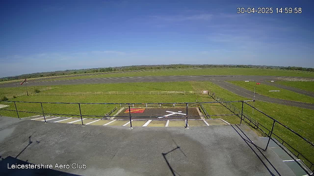 A view from a webcam at Leicestershire Aero Club showing a clear day with blue skies and a few wispy clouds. In the foreground, there is a small parking area with designated parking spaces and a helicopter landing pad marked with an "X." Surrounding the area are green grassy fields, with a small runway or taxiway visible extending into the distance. There are fencing and safety barriers along the edges of the landing area. The timestamp in the upper right corner reads 30-04-2025 14:59:58.
