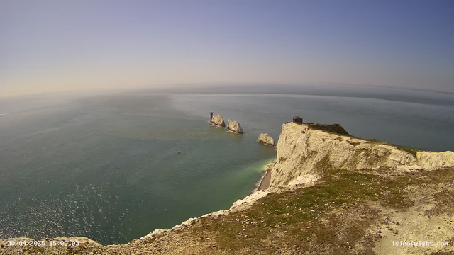 Aerial view of white chalk cliffs overlooking the sea, with a few large rock formations rising from the water's surface. A small building or structure is visible on top of the cliffs, and a narrow path runs along the edge of the cliffs. The sky is clear with a gradient from pale yellow near the horizon to blue higher up. The sea appears calm with gentle ripples, and a few boats can be seen near the rocks. The timestamp indicates the photo was taken on April 20, 2025, at 3:04 PM.
