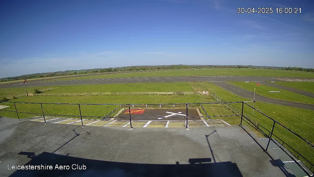 A clear, sunny day at an airfield with a blue sky and a few wispy clouds. The image shows a long, straight runway with some markings, stretching into the distance across a wide grassy area. In the foreground, there is a rooftop with a black safety railing, and a small designated helicopter landing pad marked with a white "H" and a red square, surrounded by an open grassy expanse. The timestamp in the top right corner reads "30-04-2025 16:00:21," and the bottom left corner has the text "Leicestershire Aero Club."