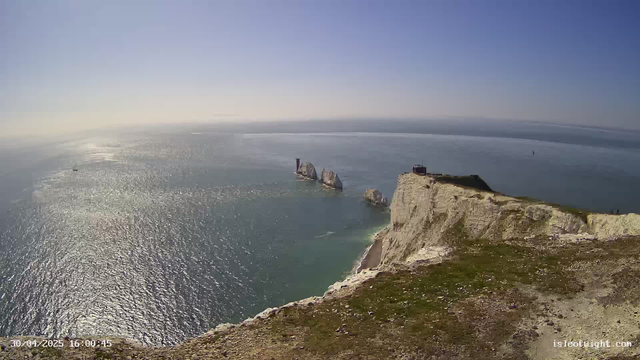 A high-angle view of white chalk cliffs overlooking the ocean on a bright, sunny day. In the distance, there are several sea stacks rising from the water, along with a few sailboats. The sky is clear and blue, and the sunlight reflects off the sea surface. The timestamp shows the date as April 30, 2025, at 16:00:45.