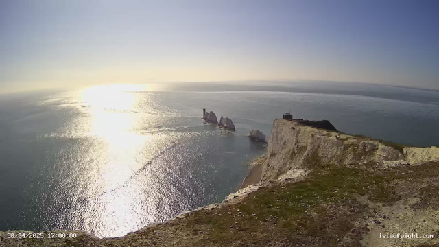 A view from a webcam showing a rocky coastline with white cliffs and a lighthouse on top. The sun is low in the sky, reflecting brightly on the calm ocean waters, creating a sparkling effect. There is green grass and dirt visible at the edge of the cliff in the foreground. The scene appears to be during the late afternoon or early evening.