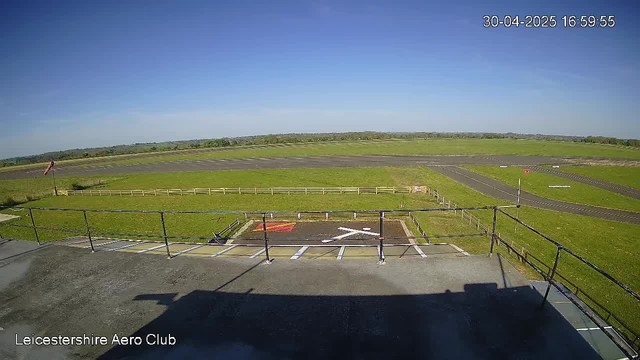 An outdoor view of a small aircraft runway at LeicesterShire Aero Club on a clear, sunny day. The foreground shows a parking area with white lines and a marked helicopter landing pad. Beyond the fence, there are grassy fields and a paved taxiway with a few markers and lights. The sky is bright blue with no clouds. The timestamp in the top right corner indicates it is 16:59:55 on April 30, 2025.