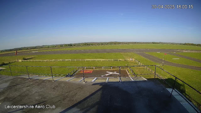 A wide open view of an airfield under a clear blue sky with some distant trees on the horizon. In the foreground, there is a platform with black railings, and the text "Leicestershire Aero Club" appears in the bottom left corner. The airstrip stretches across the middle of the image with paved taxiways and grassy areas on either side. The image is timestamped "30-04-2025 18:00:15" in the top right corner.