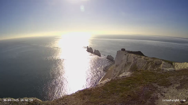 A coastal scene at sunset, showing white chalk cliffs with patches of grass on top, a small building at the edge, rock formations in the water, and the sun reflecting brightly on the calm ocean surface. The sky is clear with a gentle gradient from blue to yellow near the horizon.