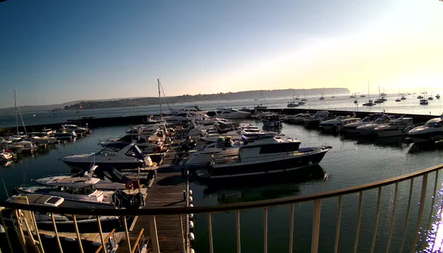A marina filled with numerous boats and yachts docked along wooden piers, with calm water reflecting the vessels. The sky is clear with a bright horizon, suggesting it is early morning or late afternoon. A railing curves around the foreground of the image.