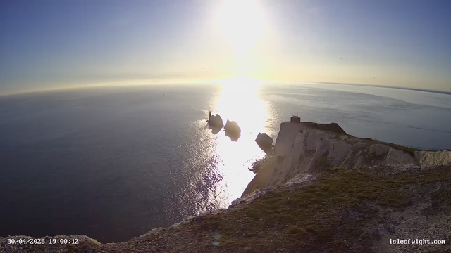 A scenic view of white chalk cliffs overlooking the ocean during sunset. The sky is clear with the sun low on the horizon, casting reflections on the water. There are a few structures on top of the cliffs, and large sea stacks are visible in the water below. The photo is taken from the edge of the cliff, with some grass and rocky terrain in the foreground.