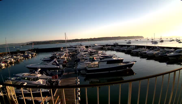 A marina filled with various boats and yachts docked at their slips, with calm water reflecting the vessels. The sky is clear, transitioning from blue to a warm yellow-orange hue near the horizon, indicating either sunrise or sunset. In the background, a distant shoreline with trees and landforms is visible.
