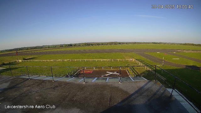 A view from a webcam shows a clear, sunny day over a flat, open landscape with grassy fields and a paved area. There is a small fenced area in the foreground, with a railing and some equipment, and a runway or track extending into the distance. The sky is mostly clear with a few wispy clouds. The date and time displayed in the top right corner are April 30, 2025, at 19:00:42. The bottom left corner has the text "Leicestershire Aero Club."