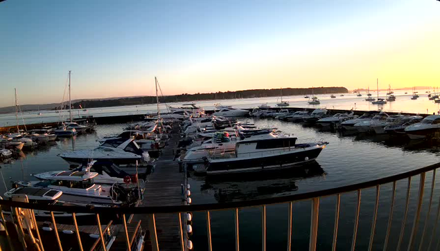 A view of a marina at sunset with several boats docked along wooden piers, and calm water reflecting the sky and boats. The horizon shows a distant shoreline, and the sky is transitioning from blue to warm sunset colors. A black metal railing is visible in the foreground.