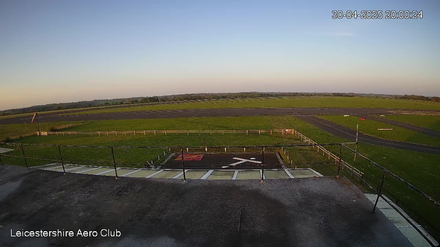 An empty helicopter landing pad with a white arrow pointing forward and a white "H" in the center, surrounded by a black fence. In the background, there are open fields with green grass and some distant trees, under a clear sky. The image has a timestamp in the top right corner showing "30-04-2025 20:00:24" and the bottom left corner displays "Leicestershire Aero Club."