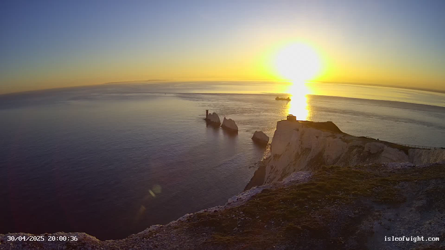 A sunset over the ocean with white chalk cliffs in the foreground. The sun is near the horizon, casting a golden reflection on the water. A pathway runs along the edge of the cliffs, and two large rock formations are visible in the water close to the shore. A ship can be seen sailing in the distance. The scene has a peaceful and scenic atmosphere.