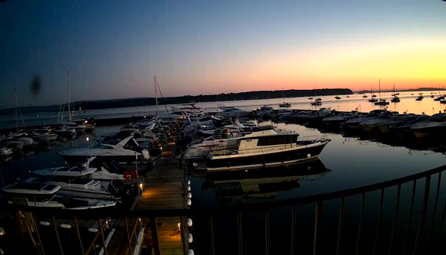 A marina filled with various boats and yachts docked at the piers during sunset. The sky is a gradient of colors from deep blue to orange and pink, and the water reflects the boats and the fading light. A black railing is visible in the foreground.