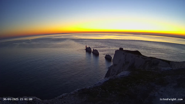A panoramic view of a coastal landscape at sunset, with sky transitioning from bright yellow and orange near the horizon to darker blue higher up. There are white chalk cliffs along the shoreline, with some large rocks and sea stacks extending into the water. A small lighthouse or navigational beacon is visible on one of the sea stacks. The calm ocean reflects the colors of the sky, and a narrow footpath or trail runs along the edge of the cliffs. The timestamp in the bottom left reads "30/04/2025 21:01:00" and the bottom right displays "isleofwight.com".