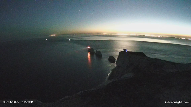 A nighttime scene of a rocky coastline with the ocean in the foreground. Several large rocks are visible emerging from the water, some equipped with small lights that cast reflections on the surface. In the distance, the horizon glows with a faint orange and yellow light, while faint city lights are visible along the shoreline. The sky is dark with a few bright stars visible. The timestamp in the lower left corner reads "30/04/2025 21:59:34," and in the lower right corner, the website "isleofwight.com" is displayed.