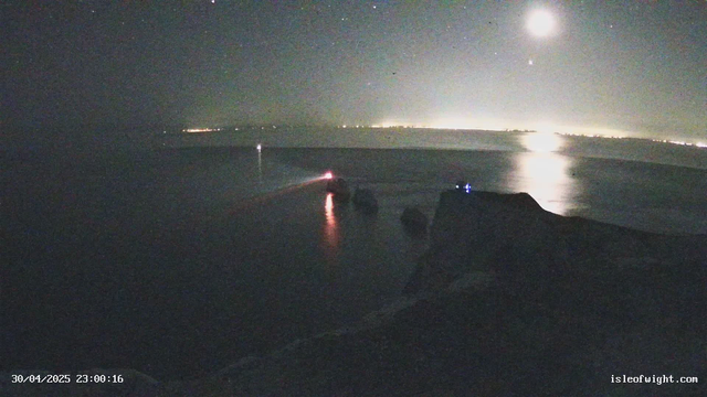 A nighttime scene of a coastal area. The sky is dark with a few visible stars, and the moon is shining brightly, casting reflections on the calm water below. There are faint lights along the horizon, possibly from distant buildings or settlements. In the foreground, there appear to be some objects or equipment on the shoreline, but they are not clearly identifiable due to low light. The timestamp in the lower left corner indicates the date and time as 30th April 2025 at 23:00.