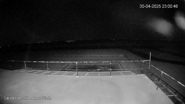 A black-and-white nighttime photo of an outdoor sports field shows empty bleachers and a fence surrounding the field. The sky is dark with a few faint stars visible, and there's a timestamp in the top right corner reading "30-04-2025 23:00:46." The bottom left corner has the text "Leicestershire Aero Club."
