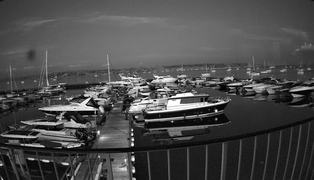 A black-and-white image of a marina with multiple boats and yachts docked along piers. The water is calm and reflects the boats, with a distant shoreline and a cloudy sky in the background. The photo is taken from a balcony or deck with a curved railing in the foreground.