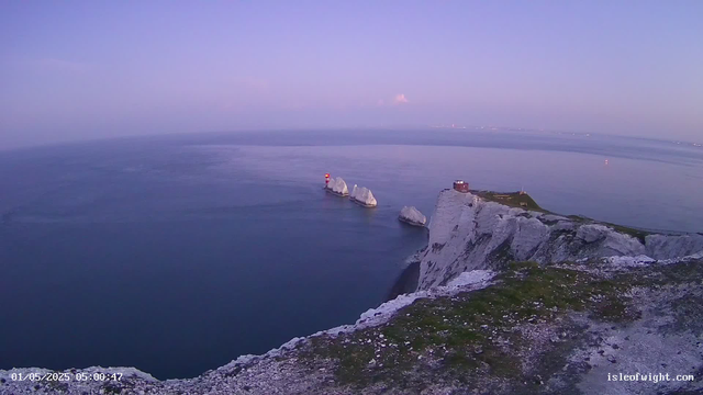 A view of white chalk cliffs along the coastline extending into calm blue ocean waters at dusk. Three rock formations with a lighthouse are situated within the sea near the cliffs. The sky is clear with a gentle gradient of soft purple and pink hues. A small building is perched on the edge of the cliffs. The timestamp in the bottom left corner indicates the date and time as May 1, 2025, at 5:00:47 AM.
