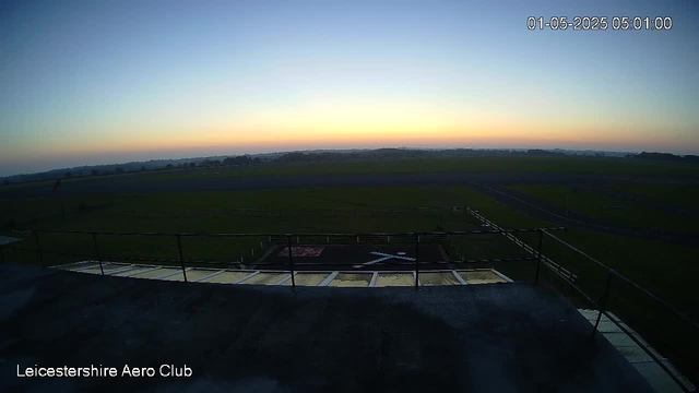 A view from a webcam shows a flat, open airfield with a helipad marked by a large white "H" in the foreground. The sky is clear with the sun either rising or setting, casting a soft light across the landscape. The horizon features distant trees and fields under a pale blue sky. The timestamp in the top right corner reads "01-05-2025 05:01:00," and the bottom left corner displays "Leicestershire Aero Club."