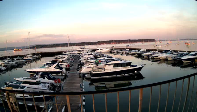 A marina with numerous boats docked along wooden piers, with calm water reflecting the boats. In the background, there is a breakwater or shoreline with more boats anchored further out. The sky is clear with a hint of sunset or sunrise, giving the scene a peaceful and serene atmosphere.