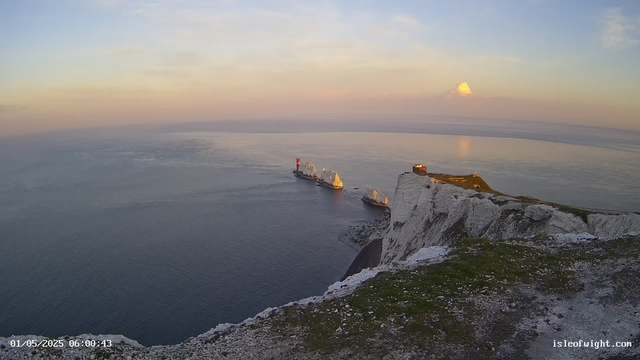 A view of white chalk cliffs along a calm sea during sunset. Several large chalk stacks, including one with a red lighthouse, stand offshore. A small building is situated on the edge of the cliffs. The sky has soft pastel colors with a few clouds, and the water reflects the warm light of the setting sun. The image appears to be taken from a webcam, with a timestamp indicating early morning.