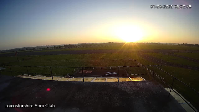 A view of an open field with a clear sky at sunrise or sunset, with the sun near the horizon casting a golden light. In the foreground, there is a concrete platform with safety railings and a small helipad marked with a white helicopter icon. The landscape appears to be at the Leicestershire Aero Club. The timestamp in the top right corner reads "01-05-2025 06:00:58."